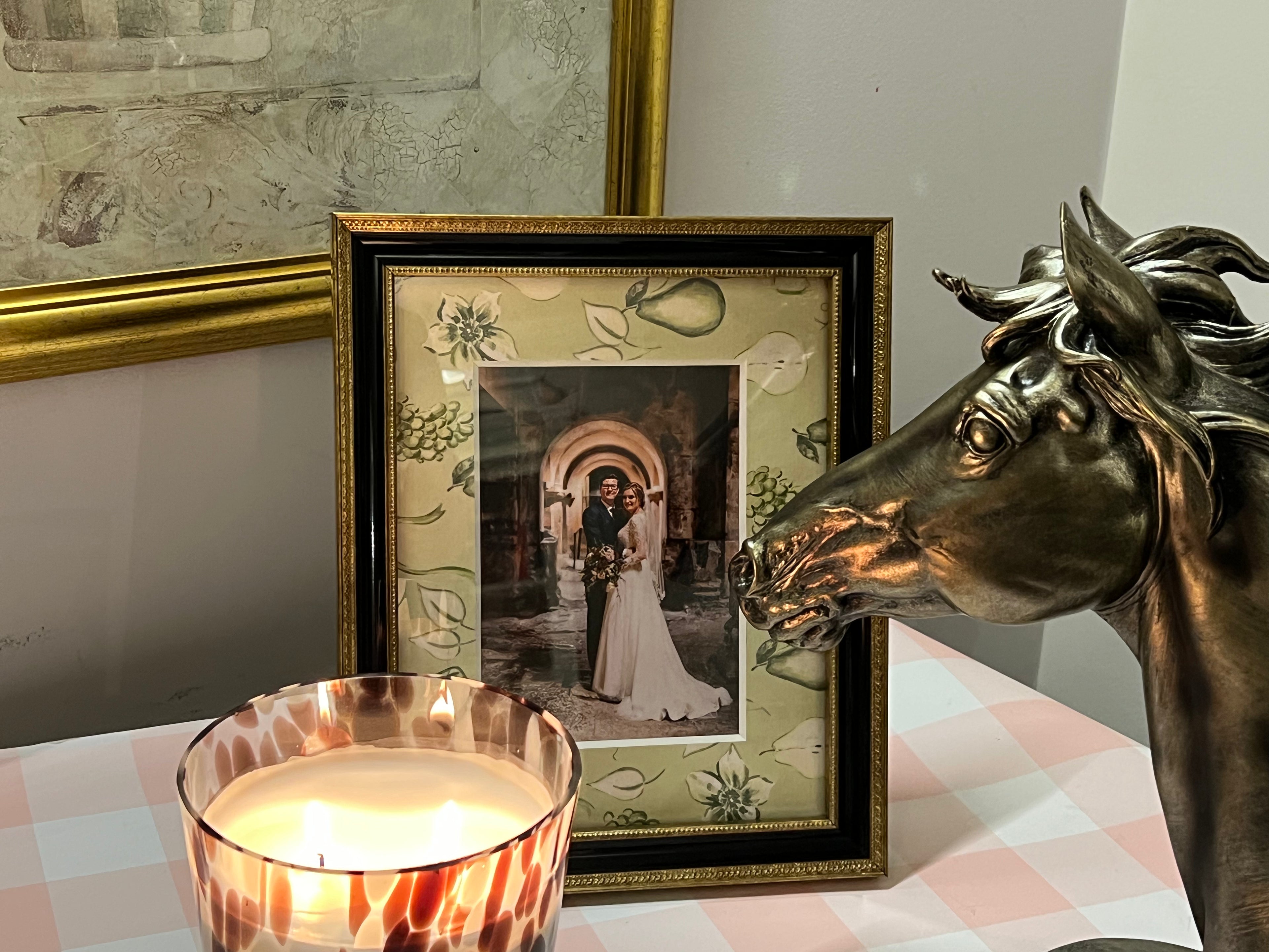 A square framed photo print featuring a bride and groom in an archway, styled on a pink-and-white checkered tablecloth next to a lit candle and a bronze horse statue.