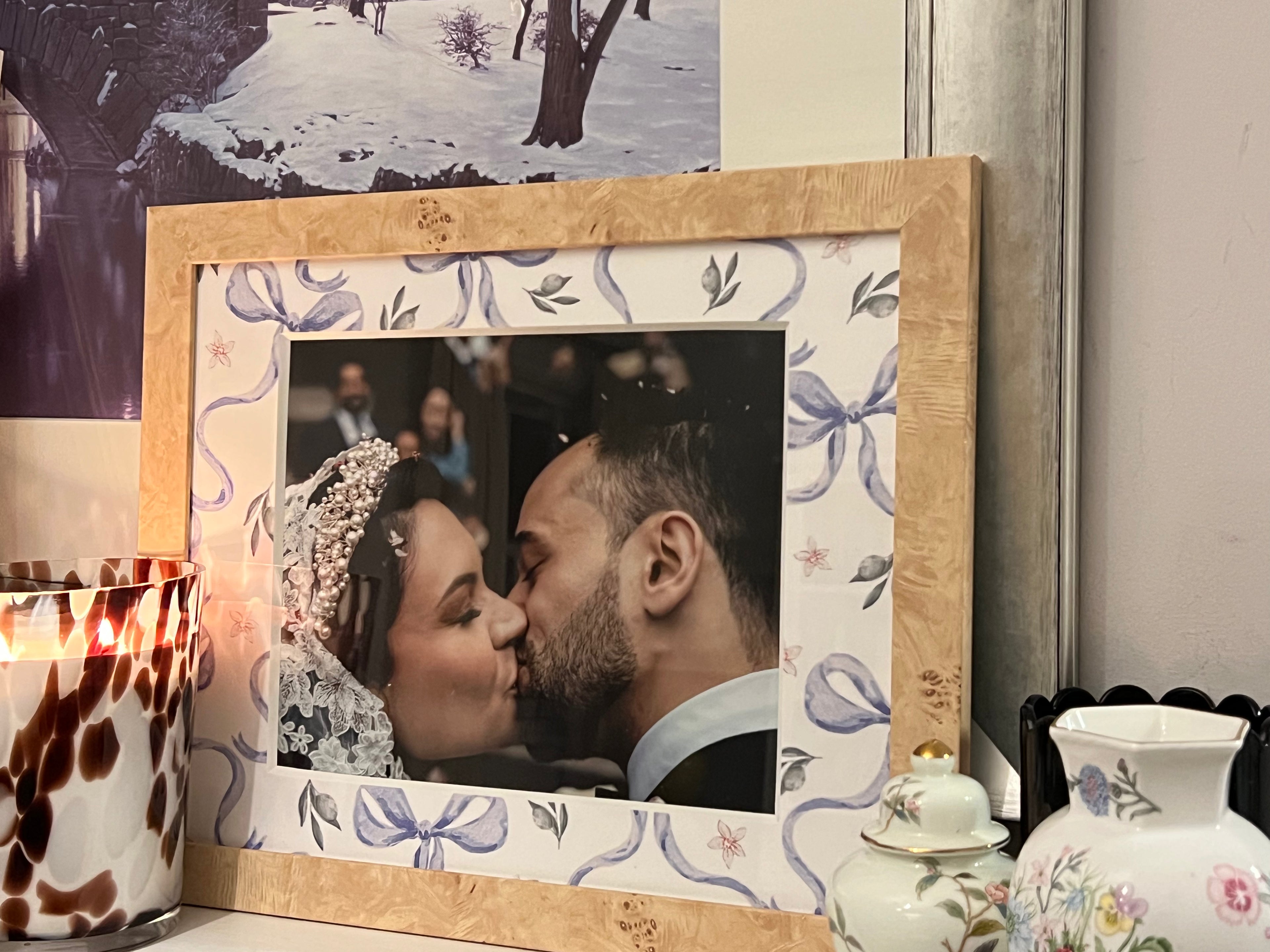 A wooden picture frame with a floral bow patterned mat holding a wedding photo of a couple kissing, styled on a tabletop next to a lit candle and ceramic vases.