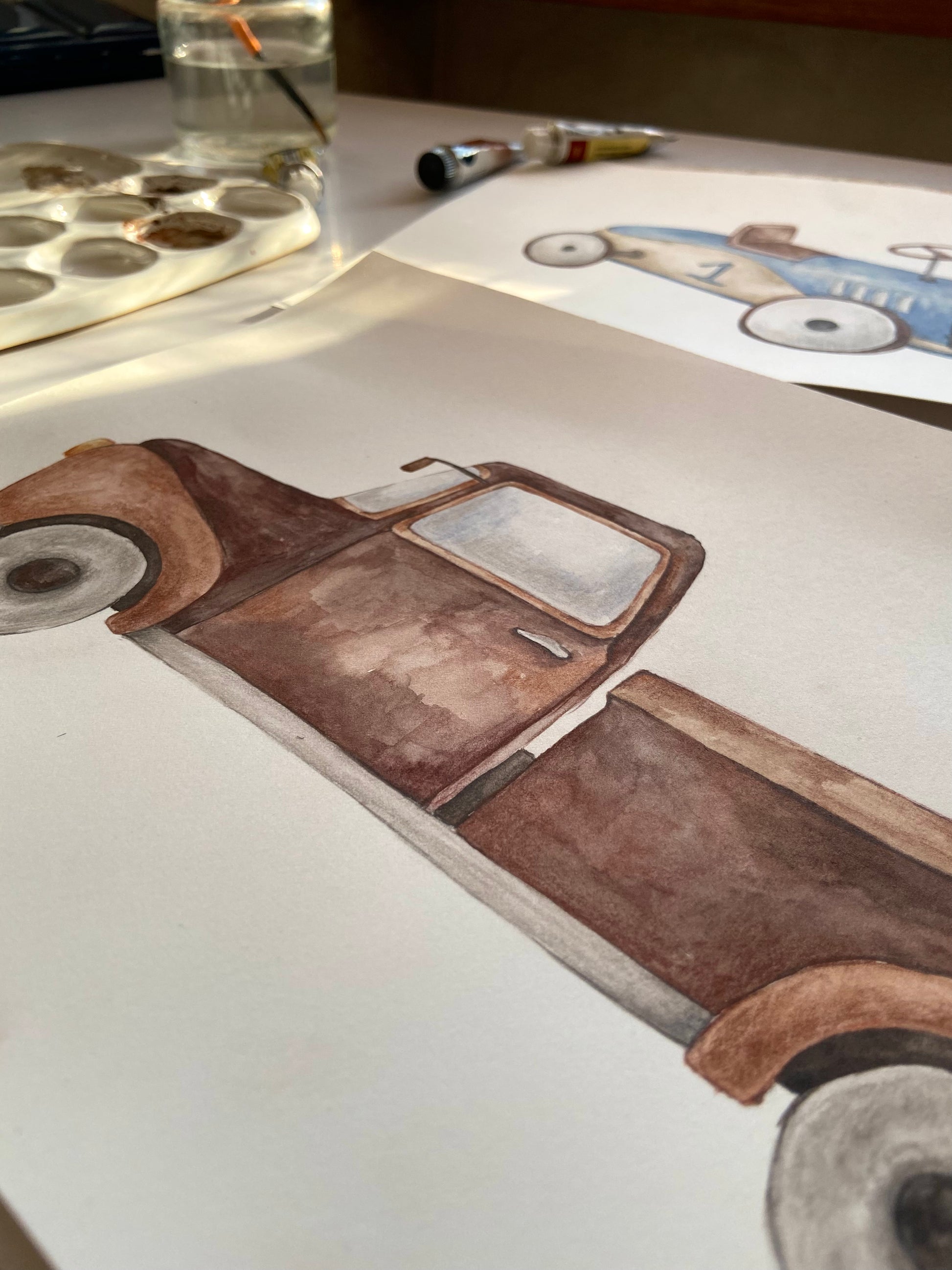 A watercolor painting of a vintage brown pickup truck on white paper, shown on a desk with paint tubes, a paint palette, and another drawing in the background.