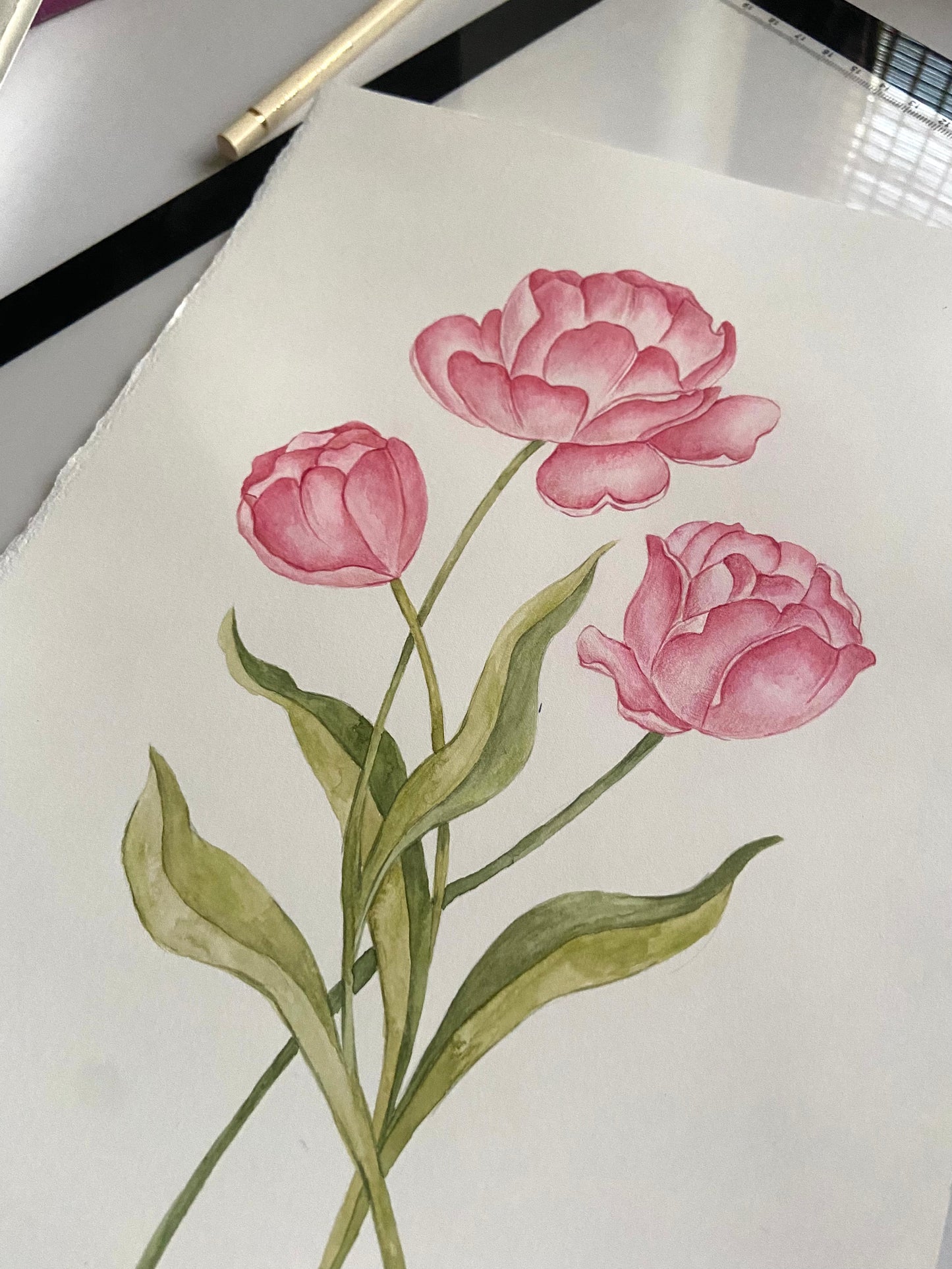 A watercolor-style floral artwork on white paper showing three pink flowers with green stems and leaves, photographed with part of a pen or pencil visible at the top edge.