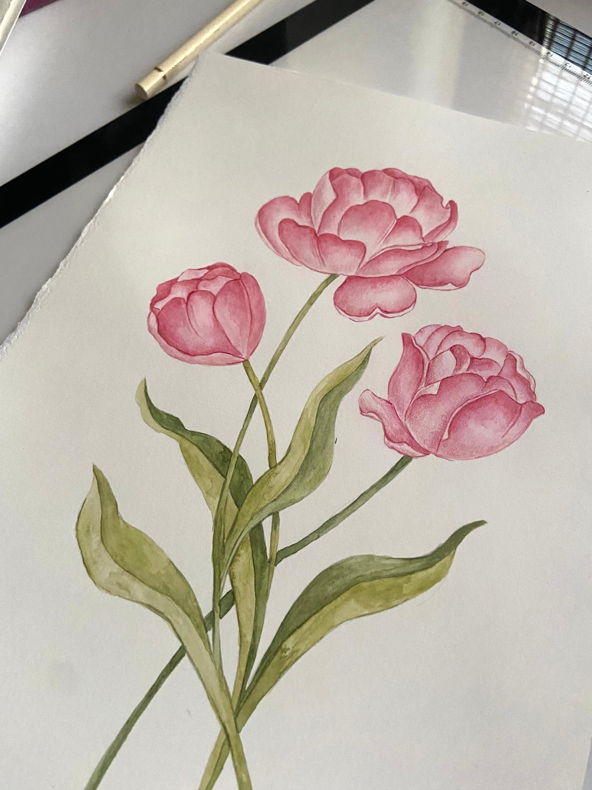 A watercolor-style floral artwork on white paper showing three pink flowers with green stems and leaves, photographed with part of a pen or pencil visible at the top edge.